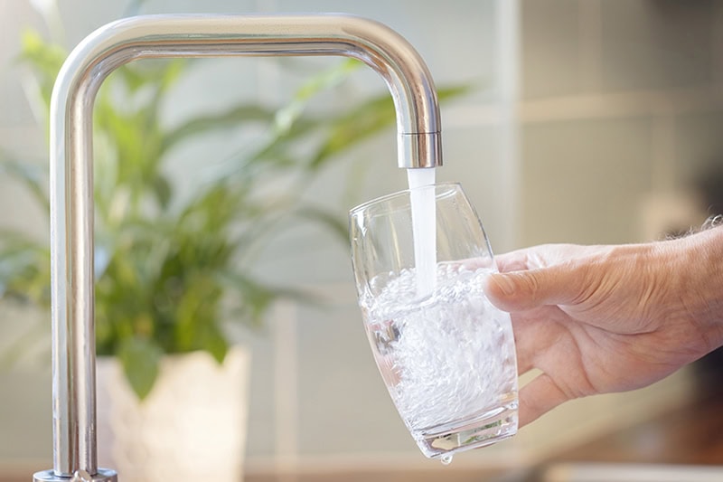 Understanding the Different Types of Water Softeners. Photo of a person filling a drinking glass of water through the kitchen faucet.